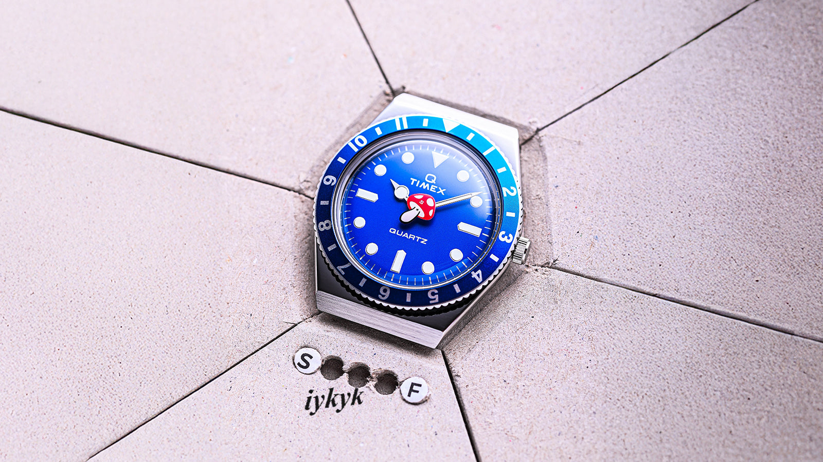 The Q Timex blue watch placed on a gray tiled surface with “iykyk” text and letters “S” and “F” arranged nearby.