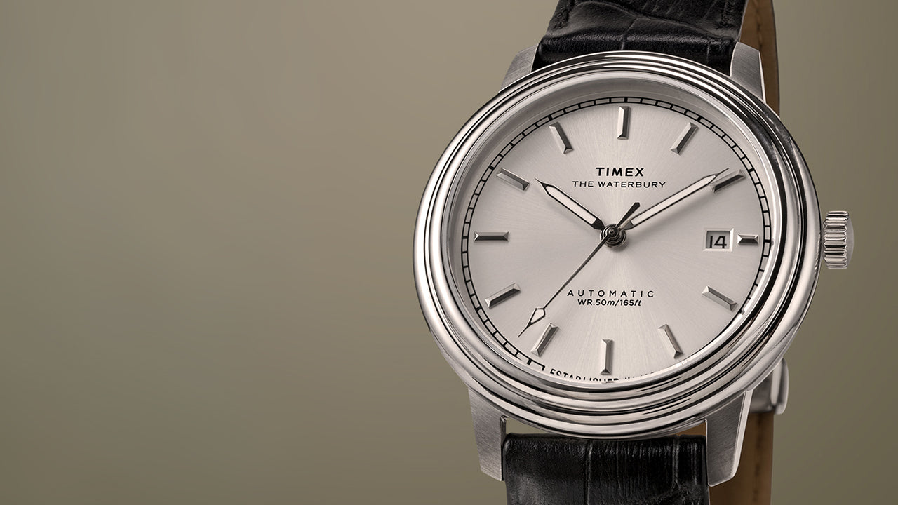 Timex Waterbury Metropolitan Automatic watch with a silver sunray dial, polished stainless steel case, and black leather strap on a beige background.