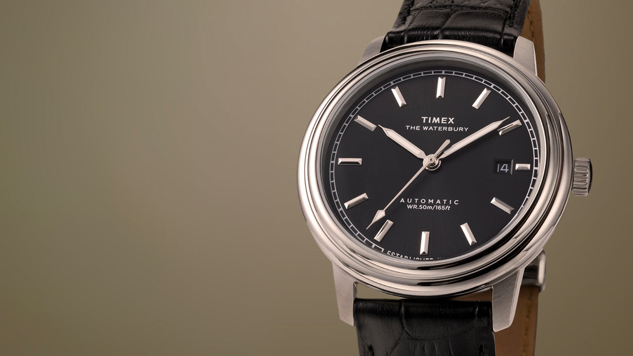 Timex Waterbury Metropolitan Automatic watch featuring a black dial, polished stainless steel case, and black leather strap on a neutral beige background.