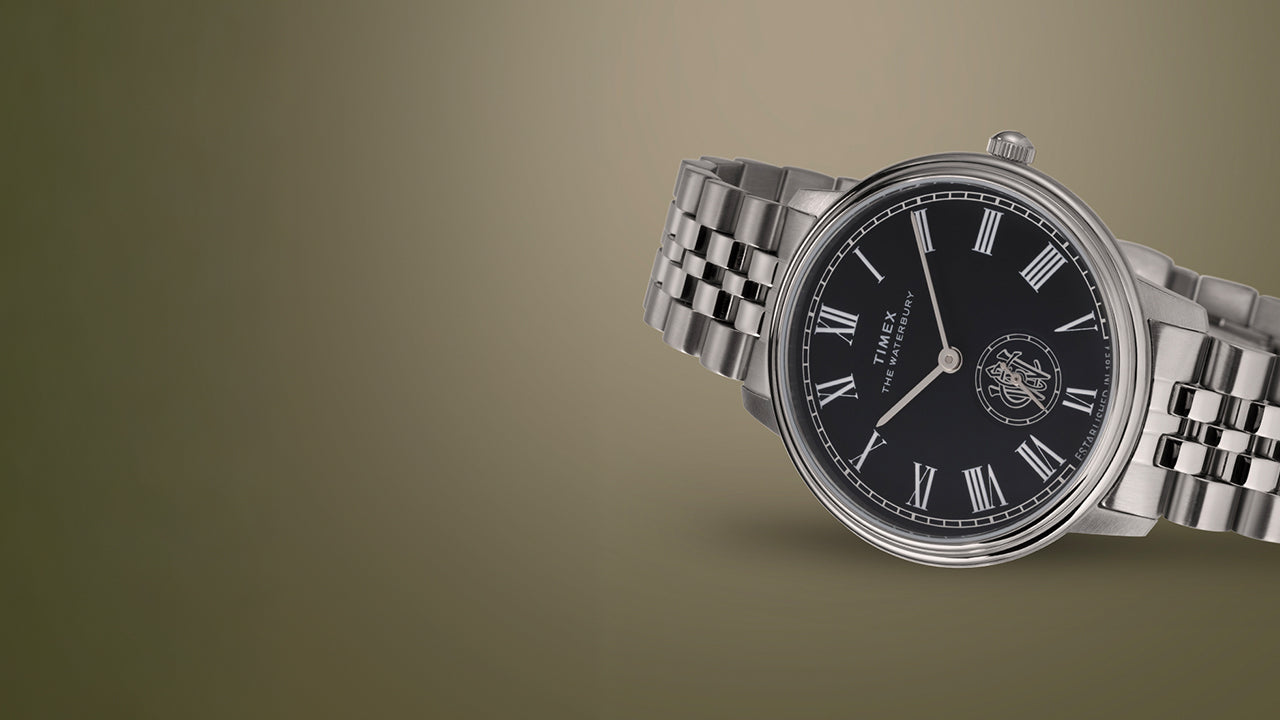 Timex Waterbury Metropolitan Sub-Second watch with polished stainless steel bracelet and black dial on a beige gradient background.