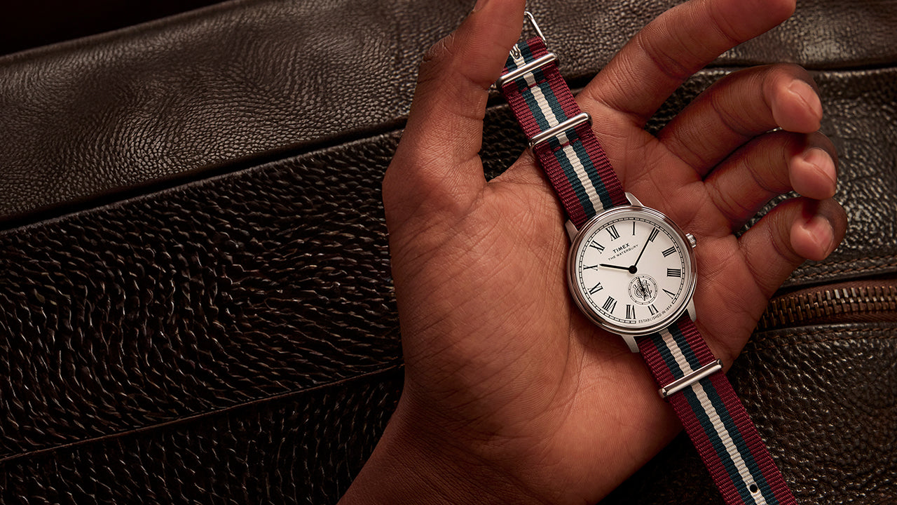 Timex Waterbury Metropolitan Sub-Second held in hand above a brown leather bag, showcasing its classic design and striped fabric strap.