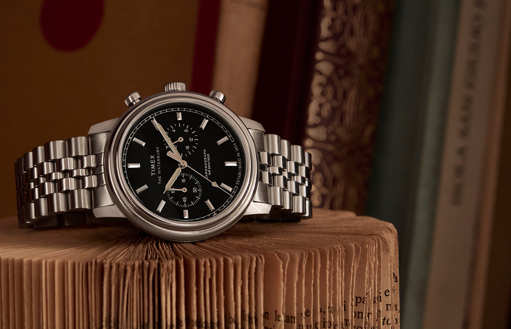 Timex Waterbury Metropolitan Chronograph with black dial and stainless steel bracelet resting on an open vintage book, showcasing its polished finish and sub-dials.