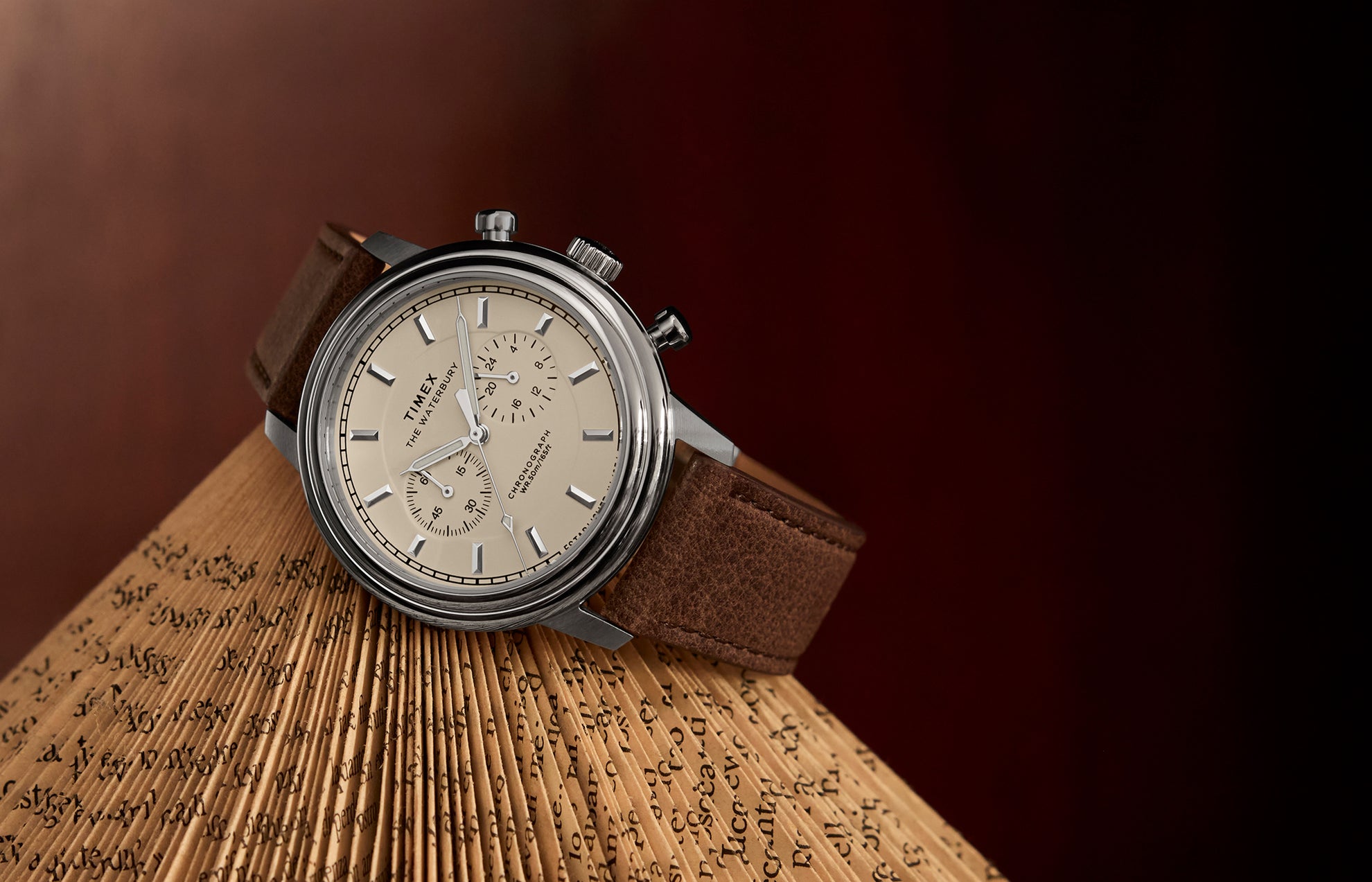 Timex Waterbury Metropolitan Chronograph with cream dial, stainless steel case, and brown leather strap resting on an open book with folded pages.