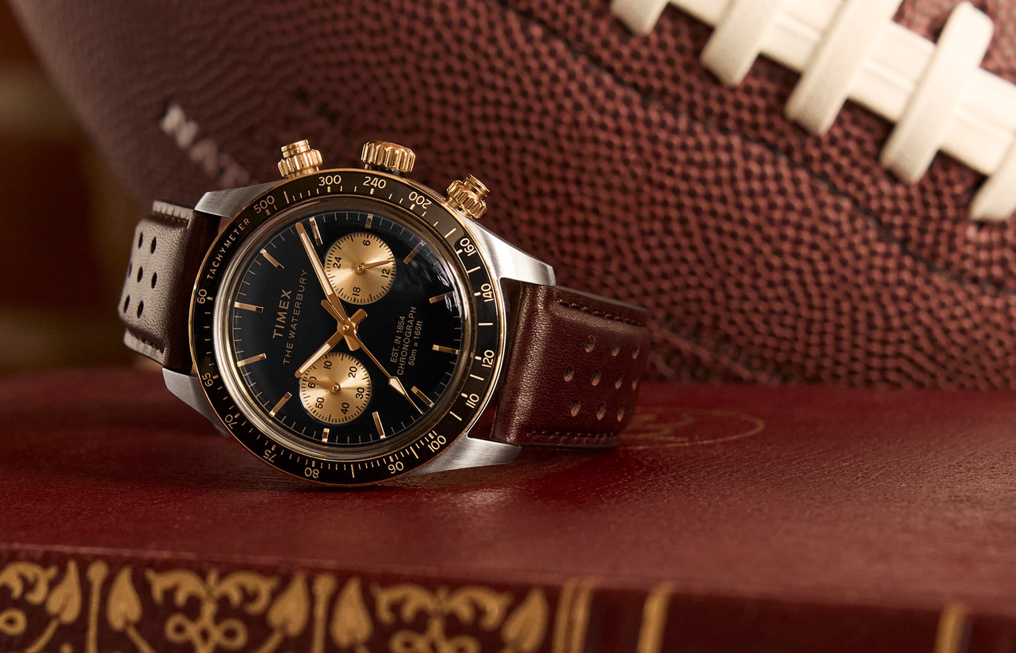 Timex Waterbury Heritage Chronograph with black dial, gold accents, and brown perforated leather strap resting on a vintage book with a football in the background.