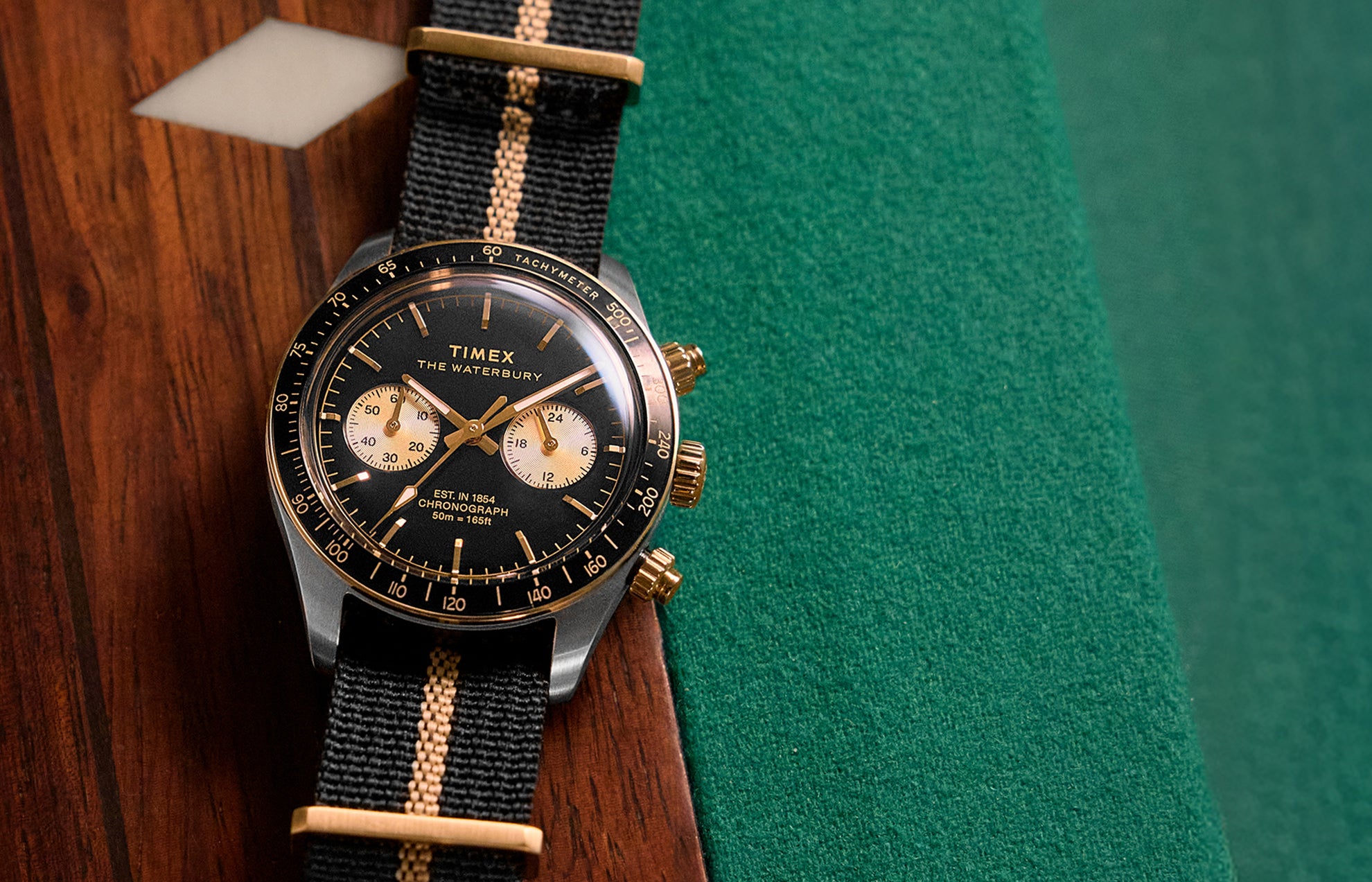 Timex Waterbury Heritage Chronograph with black dial, gold-tone accents, and black fabric strap featuring a tan center stripe resting on a wooden pool table.