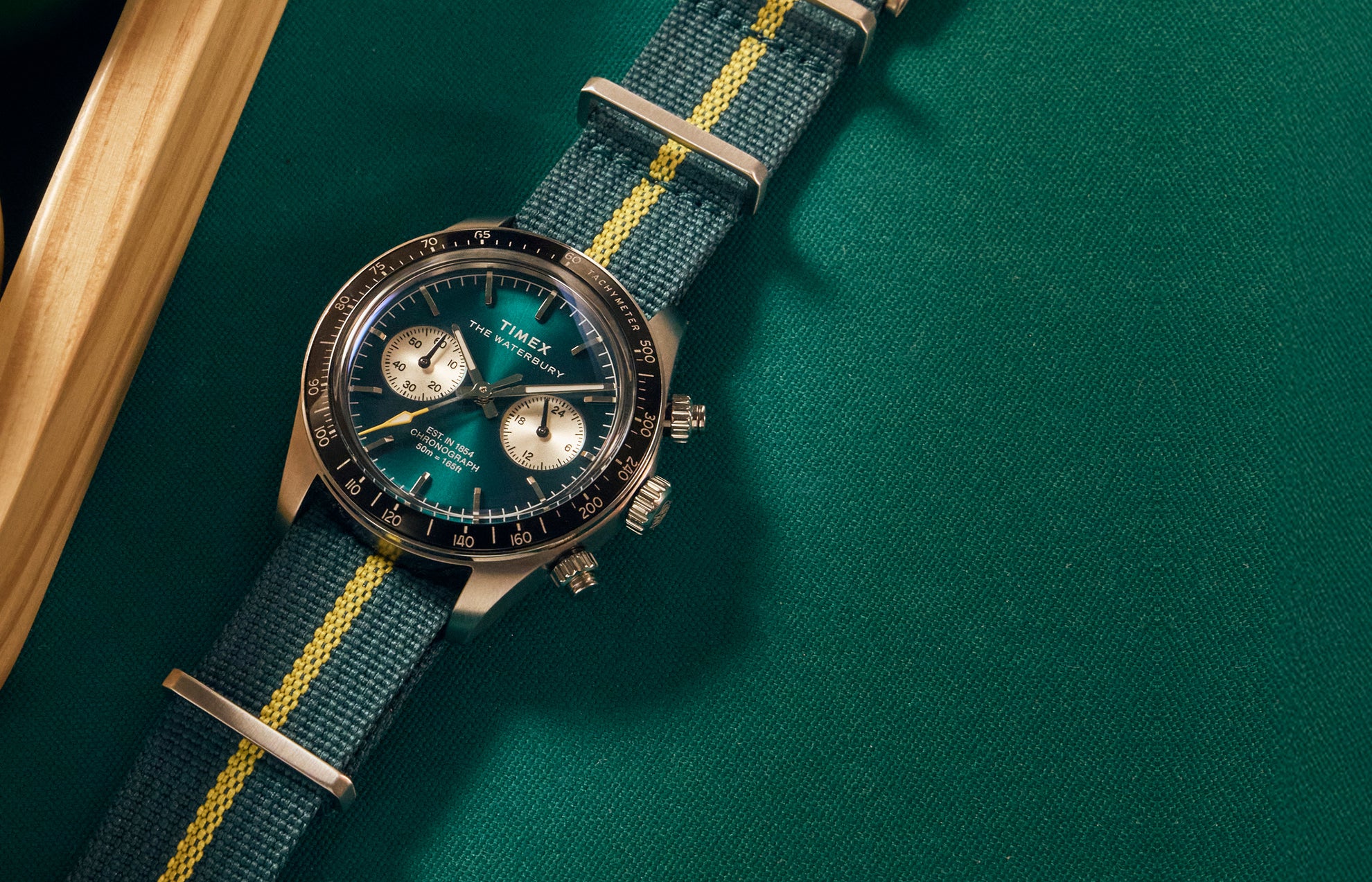 Timex Waterbury Heritage Chronograph with green sunray dial, silver sub-dials, and green fabric strap featuring a yellow center stripe resting on a billiard table.