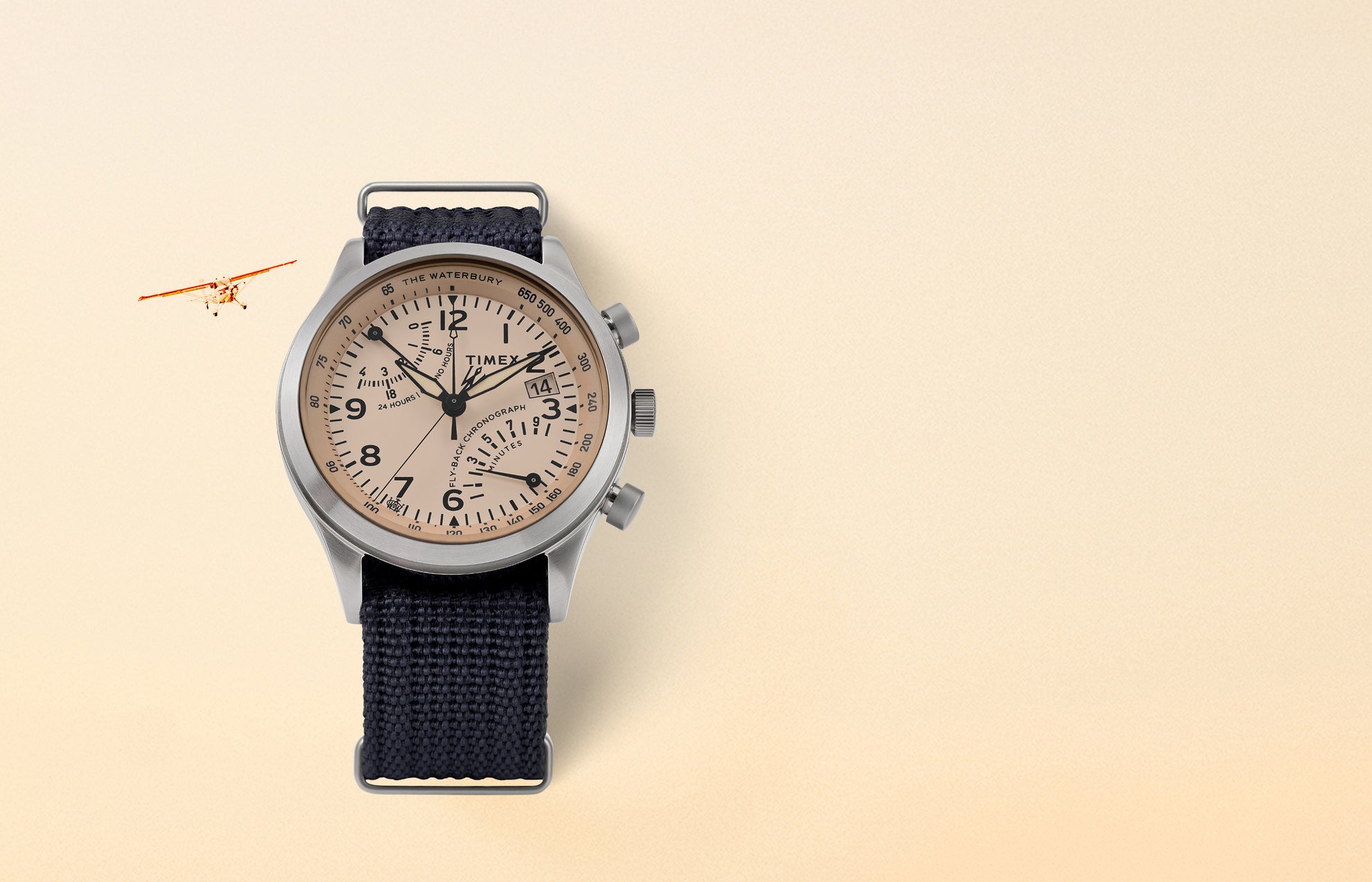 Timex Waterbury Traditional Fly-Back Chronograph with stainless-steel case, beige aviation-style dial, and navy fabric strap displayed on a cream background with a vintage plane in view.