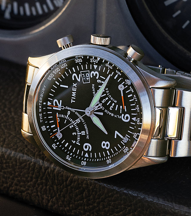 Timex Waterbury Traditional Fly-Back Chronograph resting on an aircraft instrument panel, featuring a black dial, stainless steel bracelet, and luminous hands.