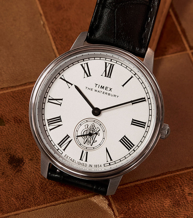 Timex Waterbury Metropolitan Sub-Second watch with white dial, Roman numerals, and black leather strap resting on a vintage chessboard.