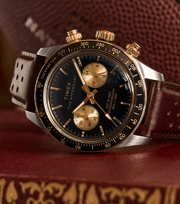 Timex Waterbury Heritage Chronograph with black dial, gold accents, and brown perforated leather strap resting on a vintage book with a football in the background.