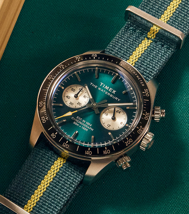 Timex Waterbury Heritage Chronograph with green sunray dial, silver sub-dials, and green fabric strap featuring a yellow center stripe resting on a billiard table.