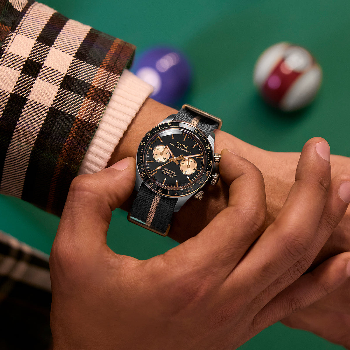 Close-up of the Timex Waterbury Heritage Chronograph on wrist, showing black and gold dial with striped fabric strap beside pool balls.