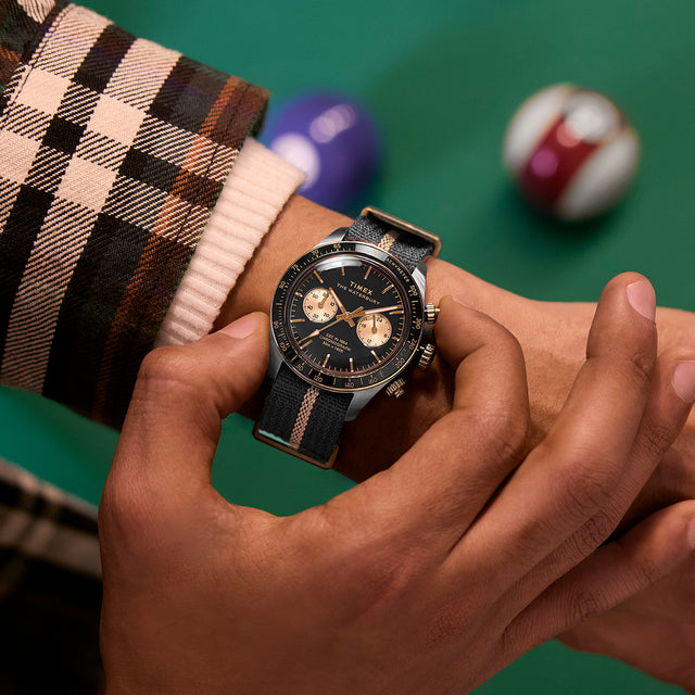 Close-up of the Timex Waterbury Heritage Chronograph on wrist, showing black and gold dial with striped fabric strap beside pool balls.
