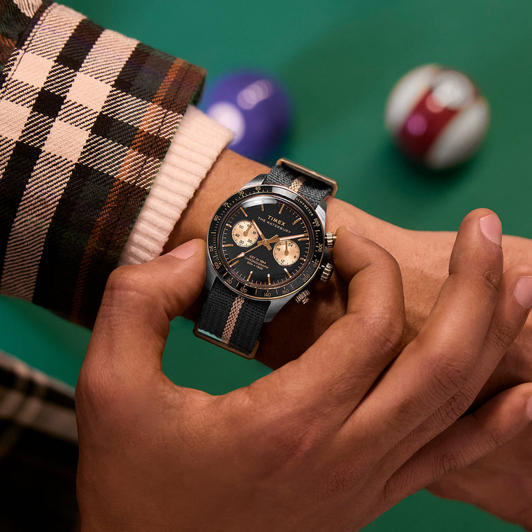 Close-up of the Timex Waterbury Heritage Chronograph on wrist, showing black and gold dial with striped fabric strap beside pool balls.
