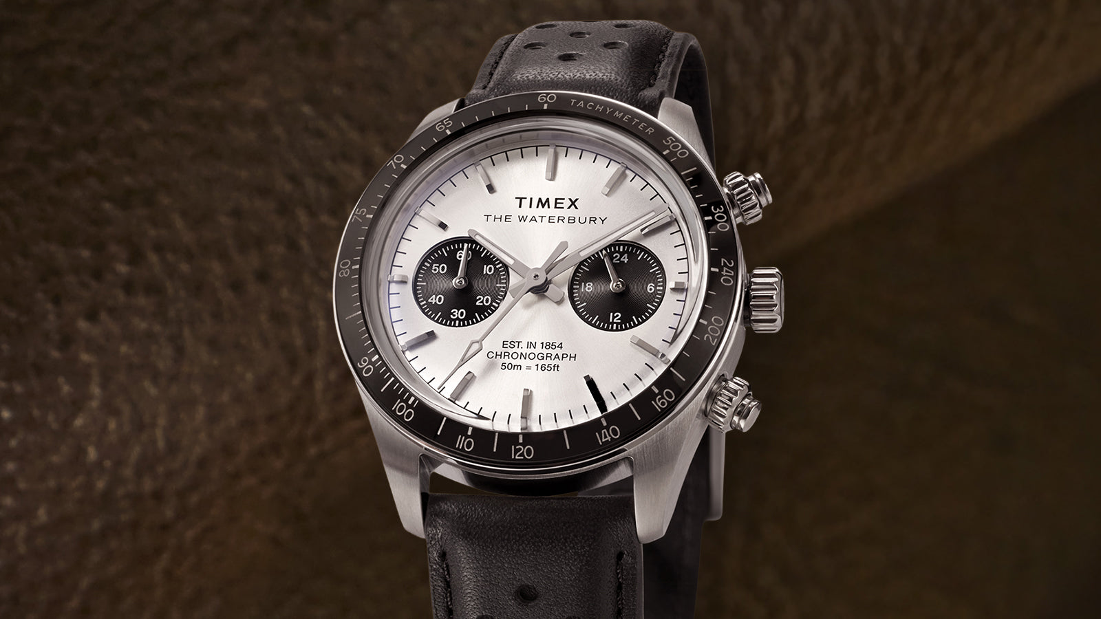 Three-quarter product view of the Timex Waterbury Heritage Chronograph with silver dial, black tachymeter bezel, and perforated leather strap.