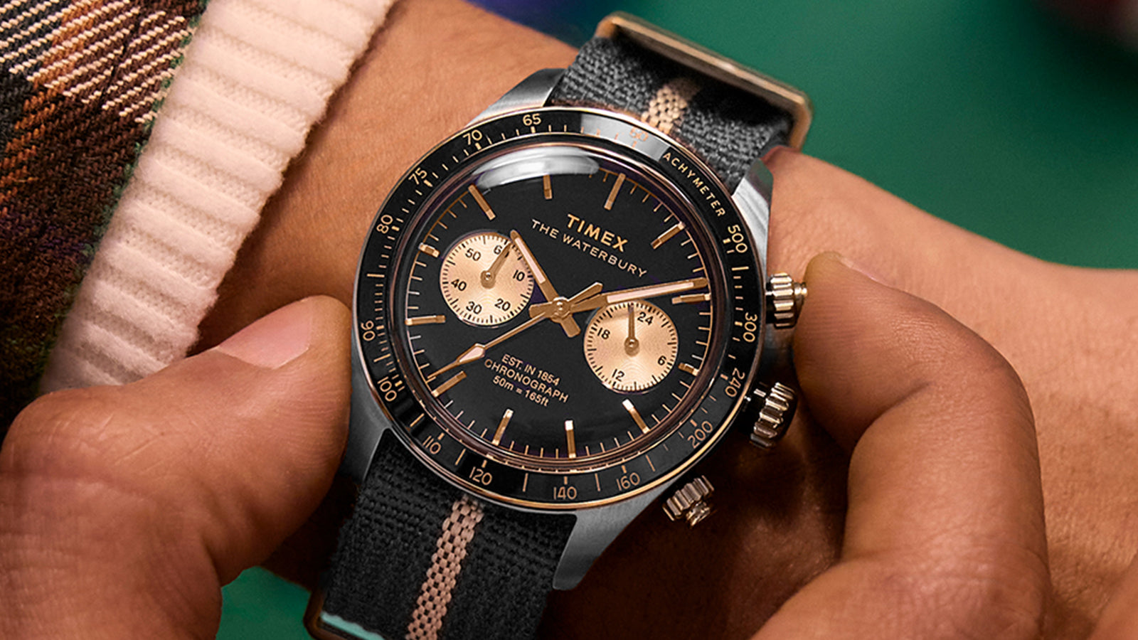 Man adjusting his Timex Waterbury Heritage Chronograph watch with black dial and gold sub-dials, paired with a striped fabric strap.