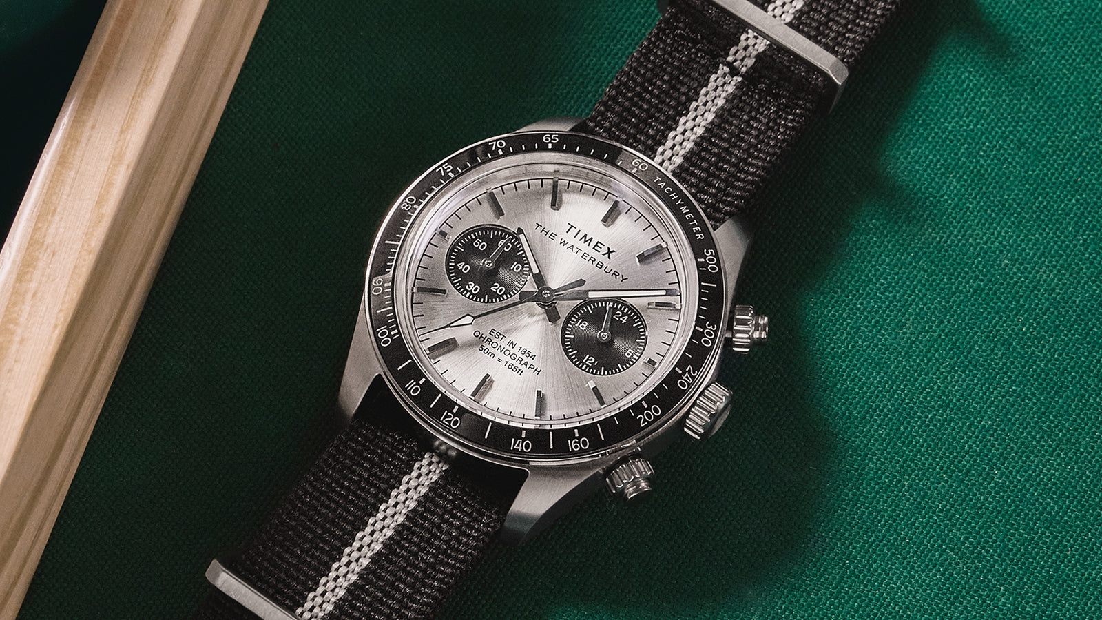 Timex Waterbury Heritage Chronograph with black and white striped fabric strap laid flat on a green pool table next to a cue stick.