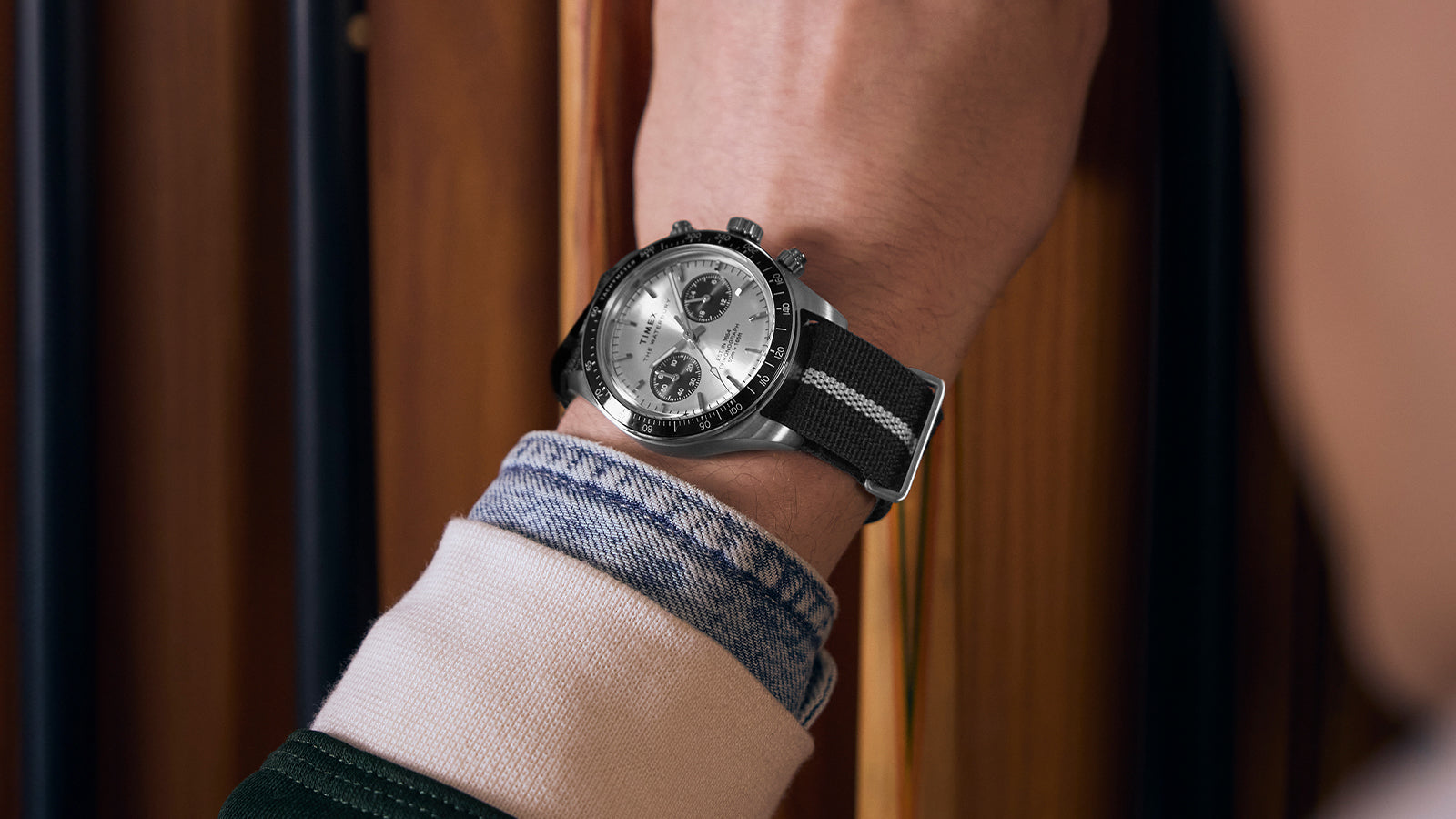 Person wearing the Timex Waterbury Heritage Chronograph with silver and black dial and striped fabric strap while holding a pool cue near a wooden rack.