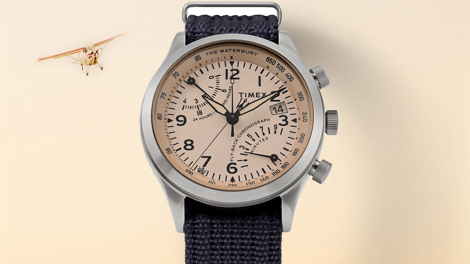 Timex Waterbury Traditional Fly-Back Chronograph with beige dial and navy fabric strap displayed against a soft cream background with a small plane flying in the distance.