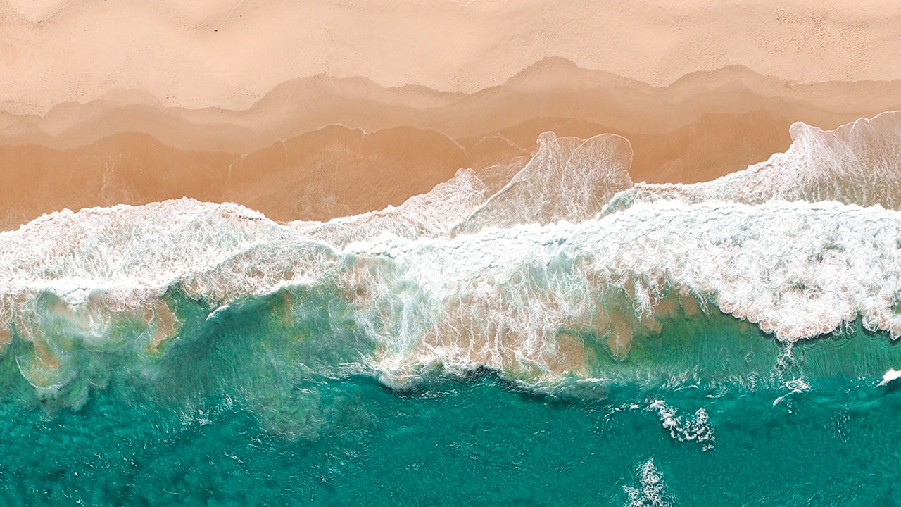 Aerial view of waves washing onto a sandy beach, symbolizing the coastal inspiration of the TIMEX® Harborside Coast collection.