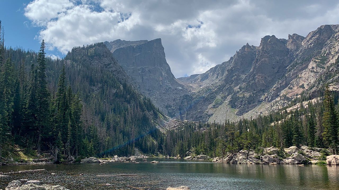 Timex Rocky Mountain National Park watch with a black case and strap, featuring a scenic mountain and forest dial design, displayed over a postcard-style background of a mountain lake.
