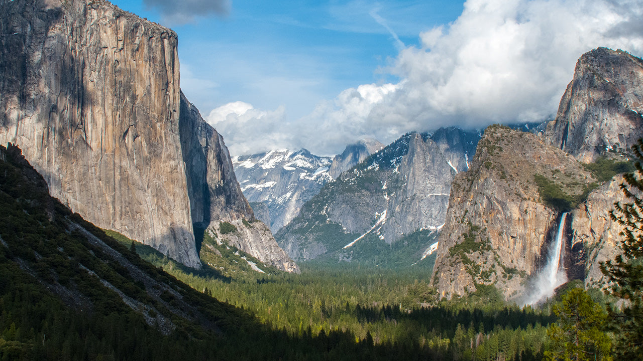 Sweeping view of a mountainous valley with cliffs, evergreen forests, and a cascading waterfall beneath a partly cloudy sky.