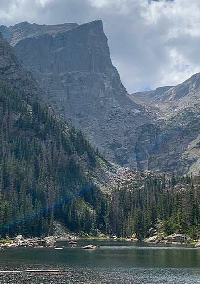 Timex Rocky Mountain National Park watch with a black case and strap, featuring a scenic mountain and forest dial design, displayed over a postcard-style background of a mountain lake.