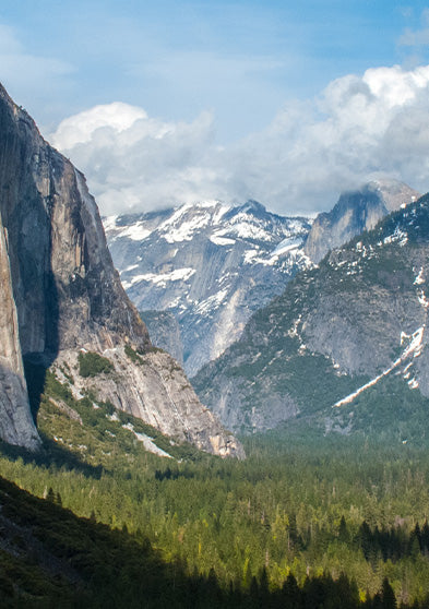 Sweeping view of a mountainous valley with cliffs, evergreen forests, and a cascading waterfall beneath a partly cloudy sky.
