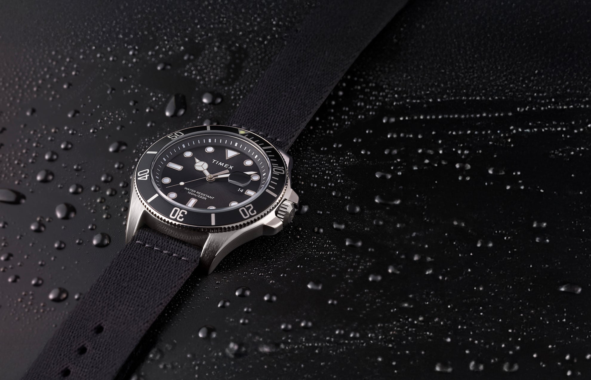 TIMEX® Harborside Coast watch with black dial and black fabric strap resting on a water-dappled black surface, showcasing its water-resistant design.