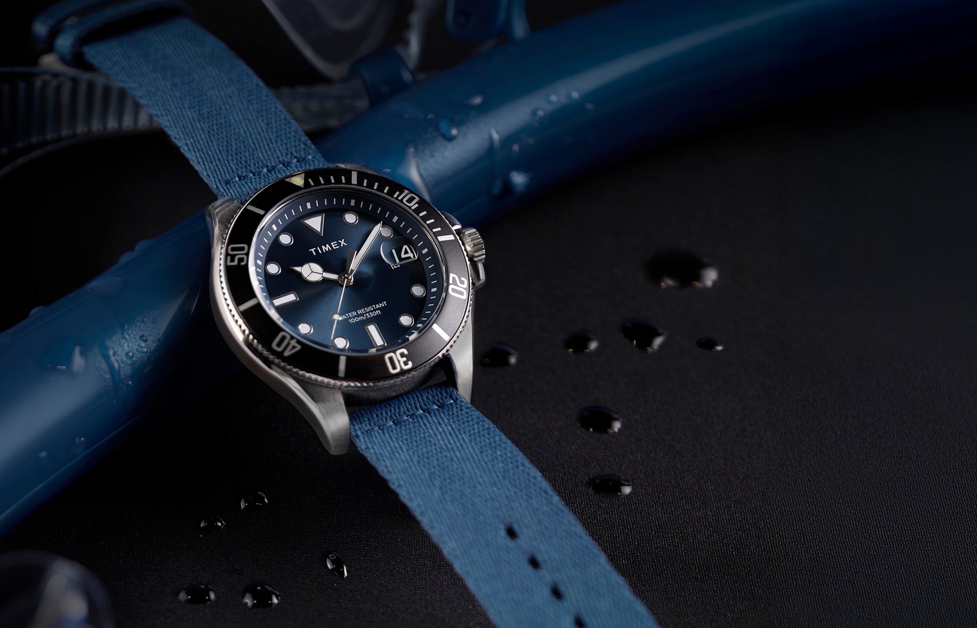 TIMEX® Harborside Coast watch with blue dial, silver-tone case, and blue fabric strap resting on a wet surface with water droplets.