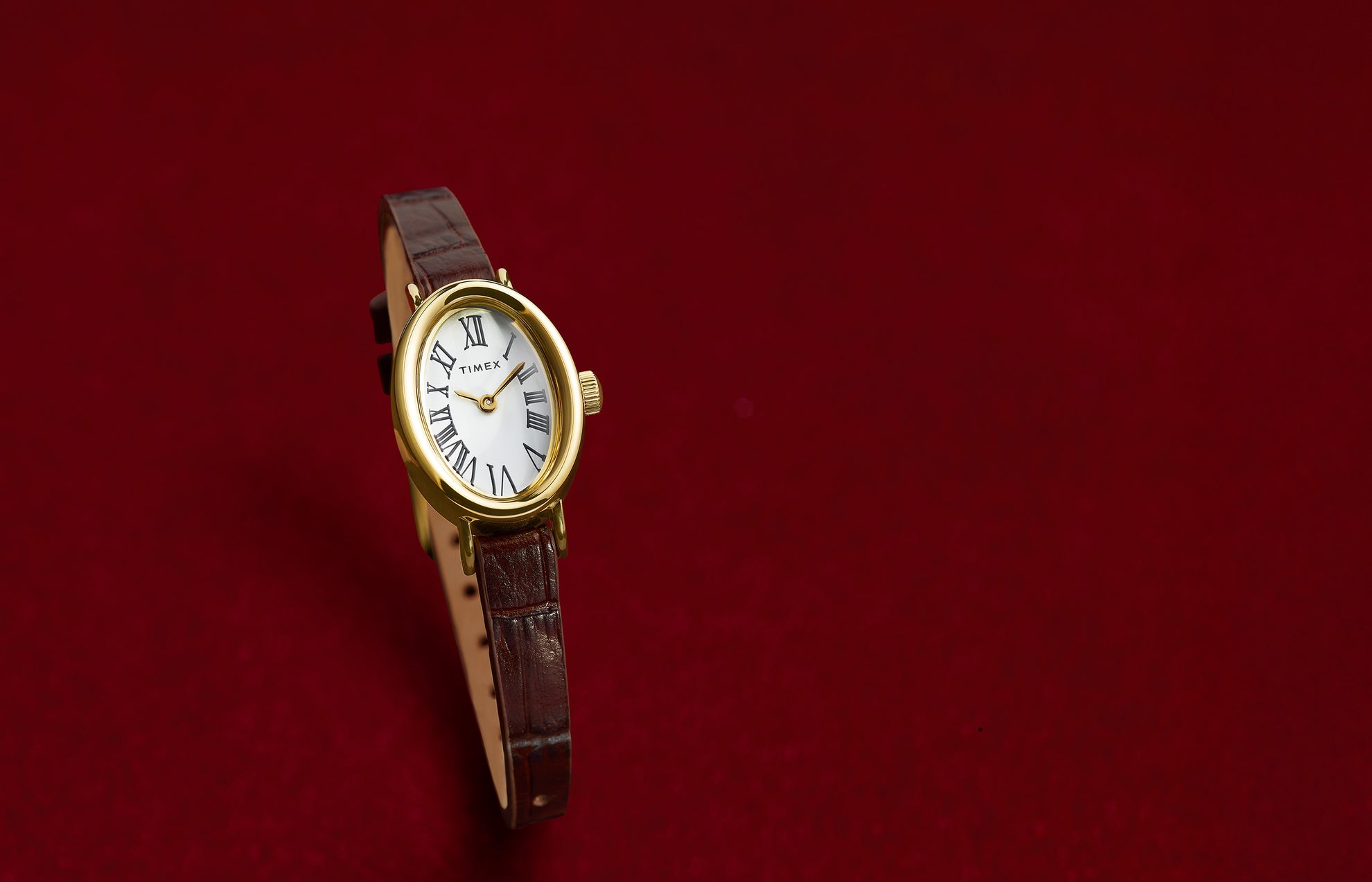 TIMEX® Cavatina watch with gold-tone case and brown leather strap displayed on a rich red background, highlighting its elegant silhouette.