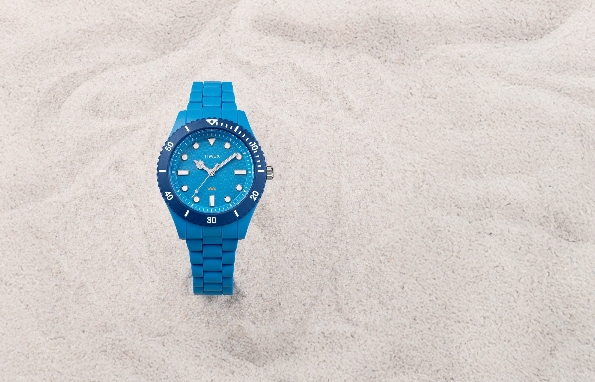 The TIMEX® Legacy Ocean #tide watch in bright blue rests on pale sand, featuring a wave-pattern dial and blue rotating bezel.