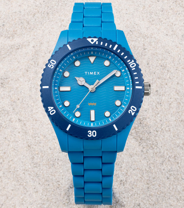 The TIMEX® Legacy Ocean #tide watch in bright blue rests on pale sand, featuring a wave-pattern dial and blue rotating bezel.