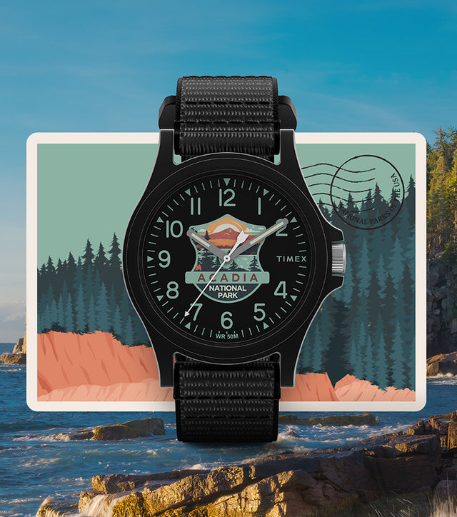 Timex Acadia National Park watch with a black case and strap, featuring a colorful mountain and forest dial design, displayed over an illustrated postcard background with a rocky coastal scene.