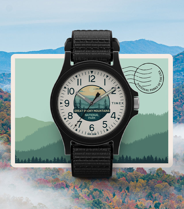 Timex Great Smoky Mountains National Park watch with black case and strap, featuring an illustrated dial of mountain scenery, displayed over a postcard-style background with a view of the Great Smoky Mountains.