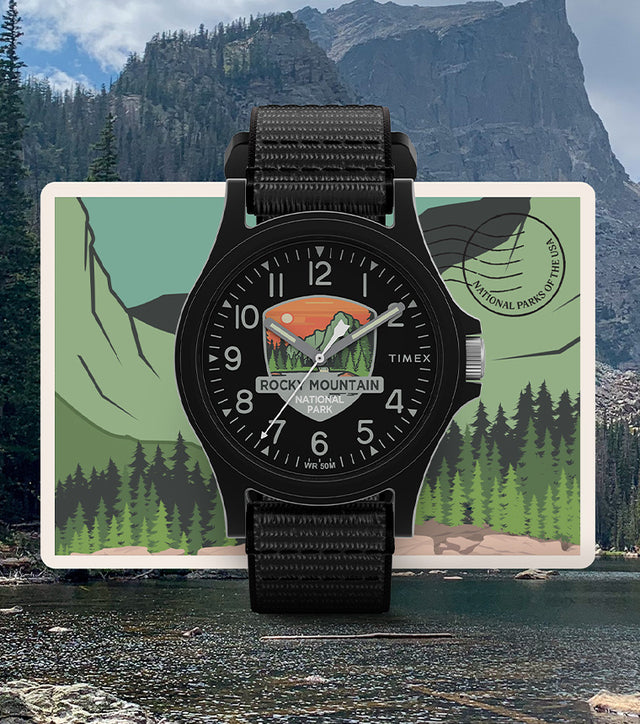 Timex Rocky Mountain National Park watch with a black case and strap, featuring a scenic mountain and forest dial design, displayed over a postcard-style background of a mountain lake.