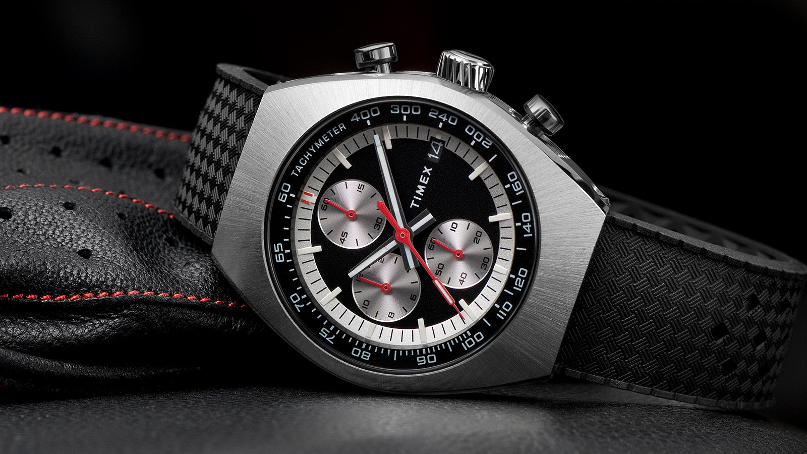 TIMEX® Legacy Tonneau Chronograph watch with black dial, silver-tone case, and textured black strap resting on perforated leather with red stitching.