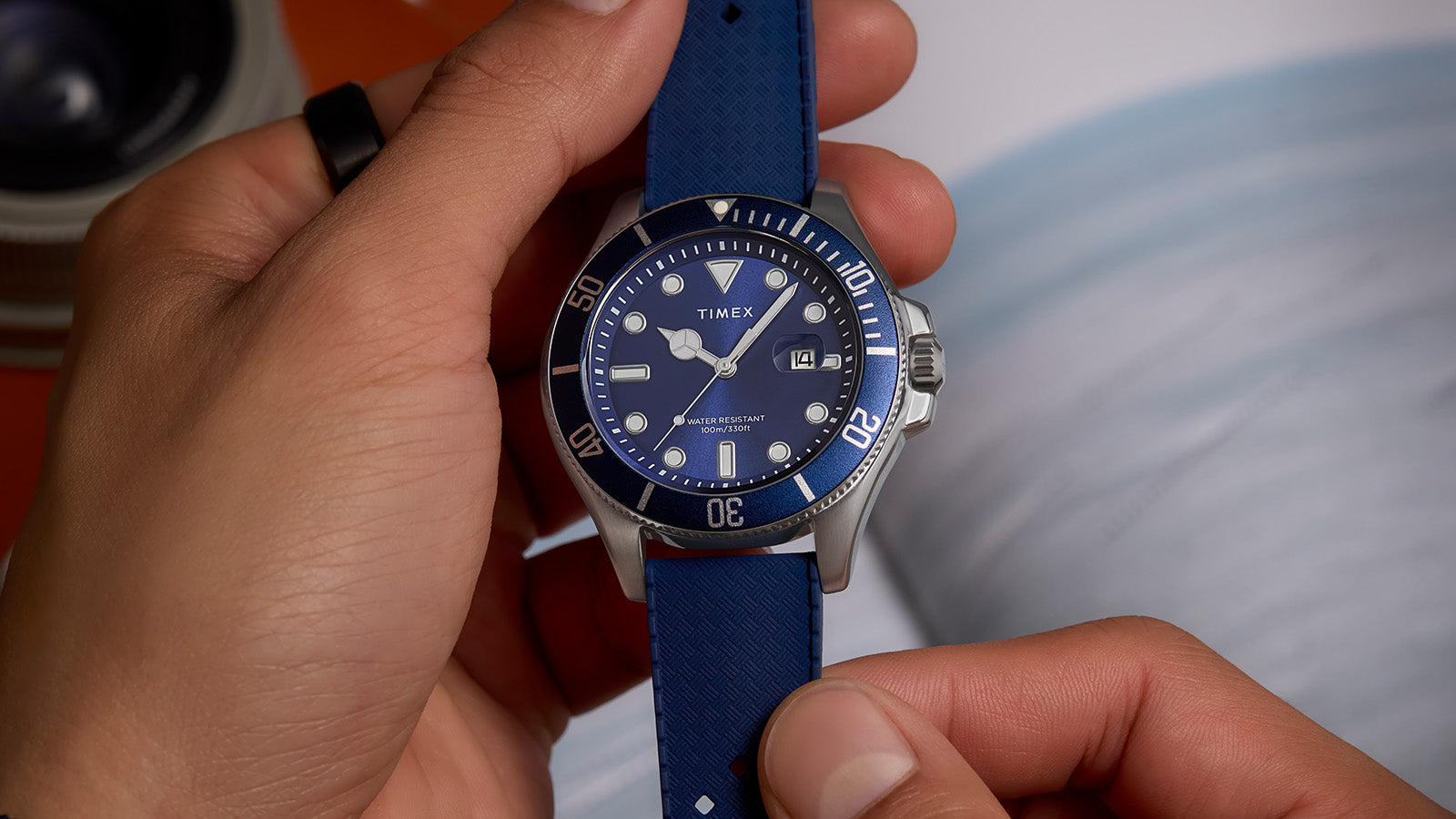 Hands adjusting a Timex Harborside Coast watch with a blue dial, blue bezel, and textured blue strap, showing date window at 3 o’clock.