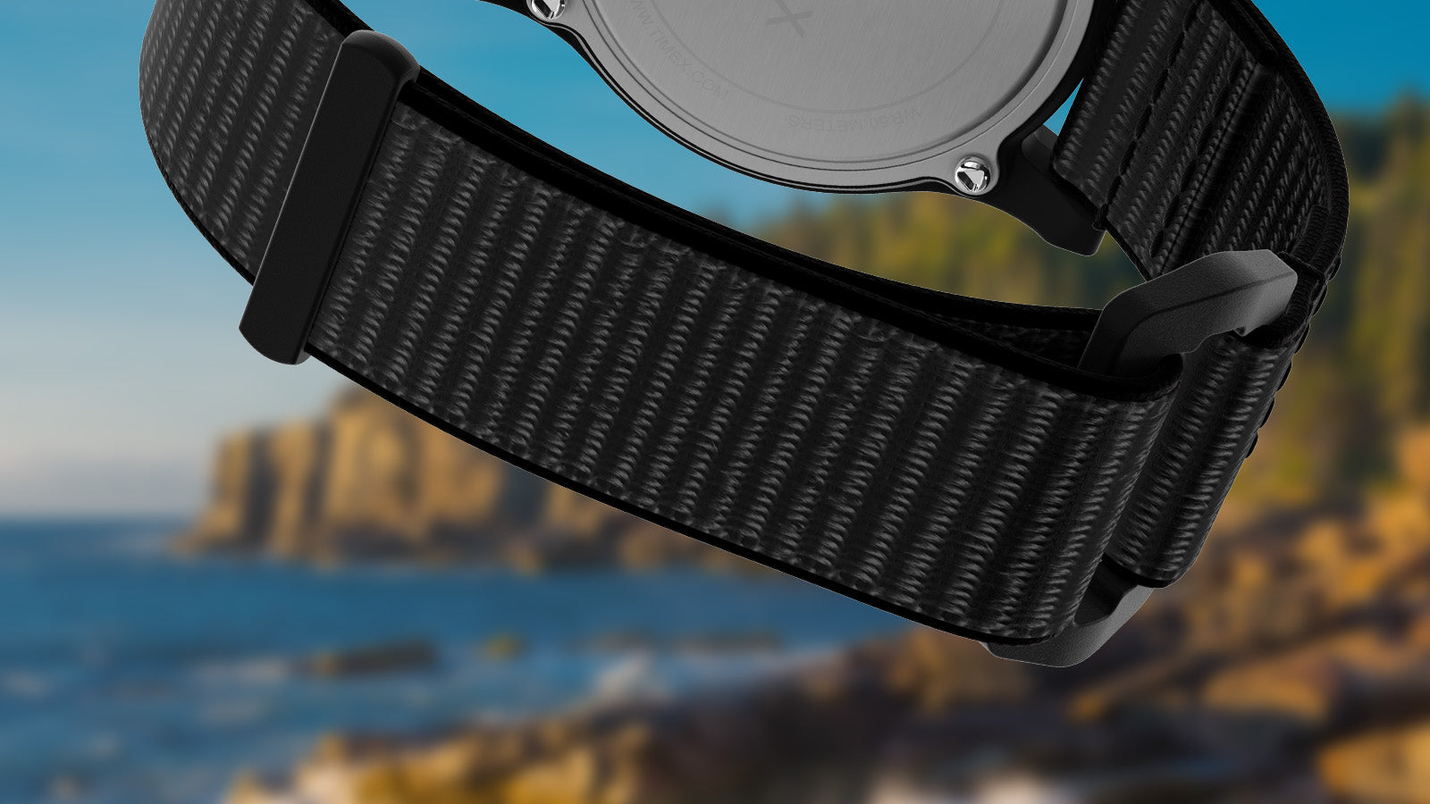 Close-up of the woven black fabric strap and buckle of the Timex Acadia National Park watch, positioned against a blurred ocean and forest background.