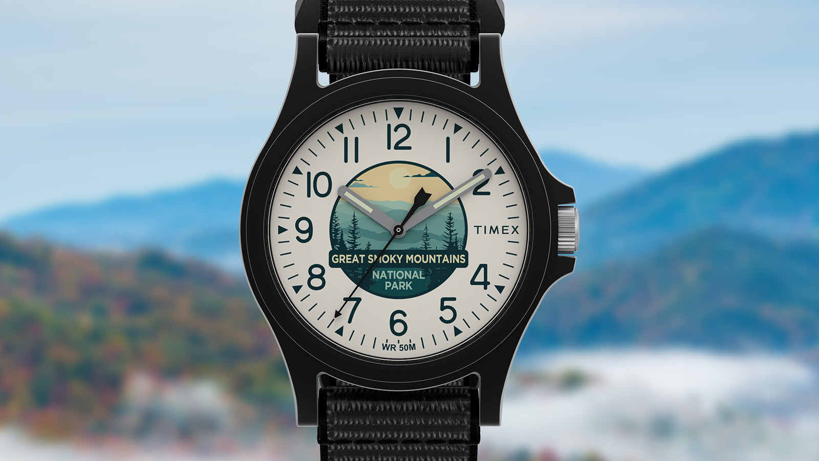 Front view of the Timex Great Smoky Mountains National Park watch with a white dial featuring mountain artwork and black nylon strap, against a soft-focus mountain landscape.
