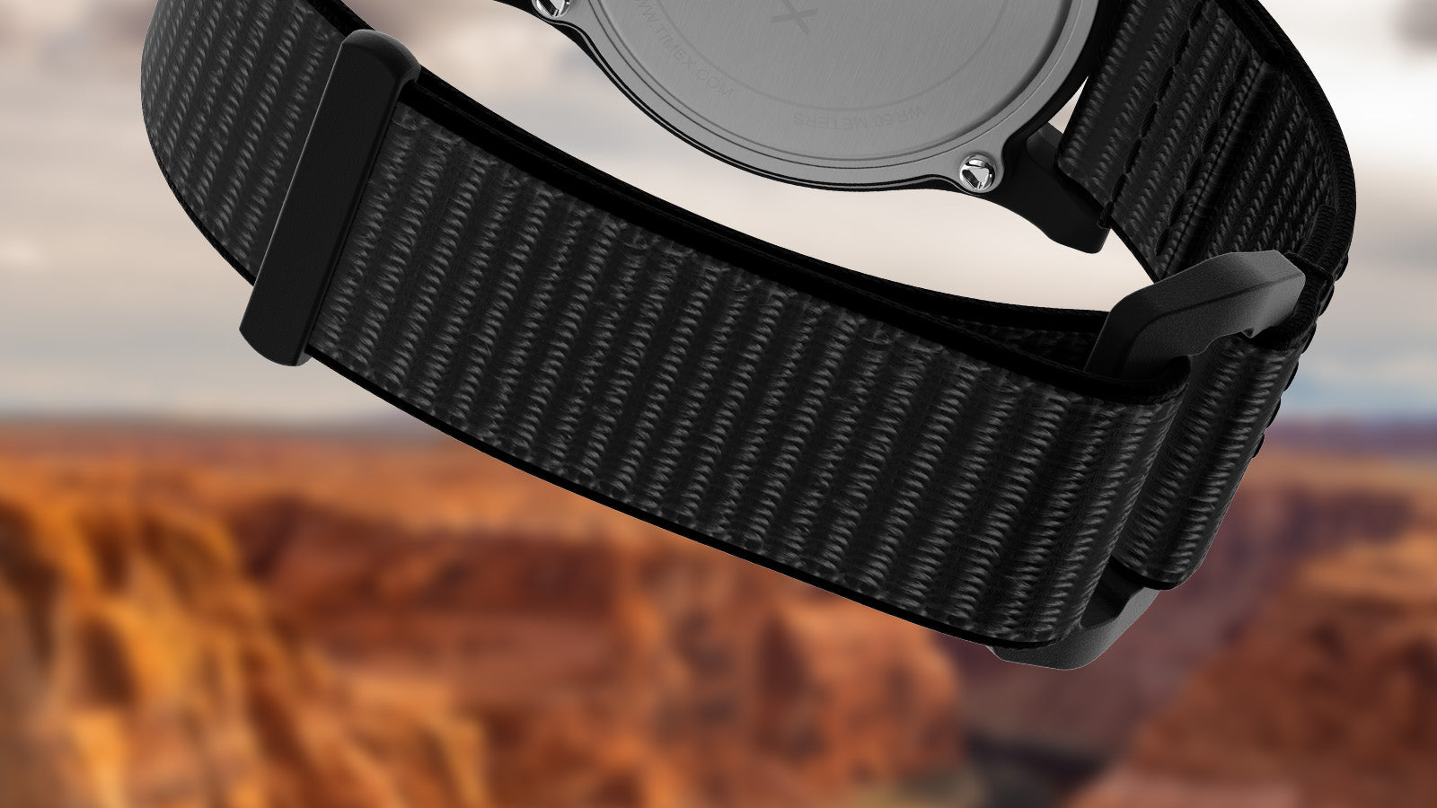 Detailed view of the black woven fabric strap and buckle of the Timex Grand Canyon National Park watch, shown against a blurred canyon background.