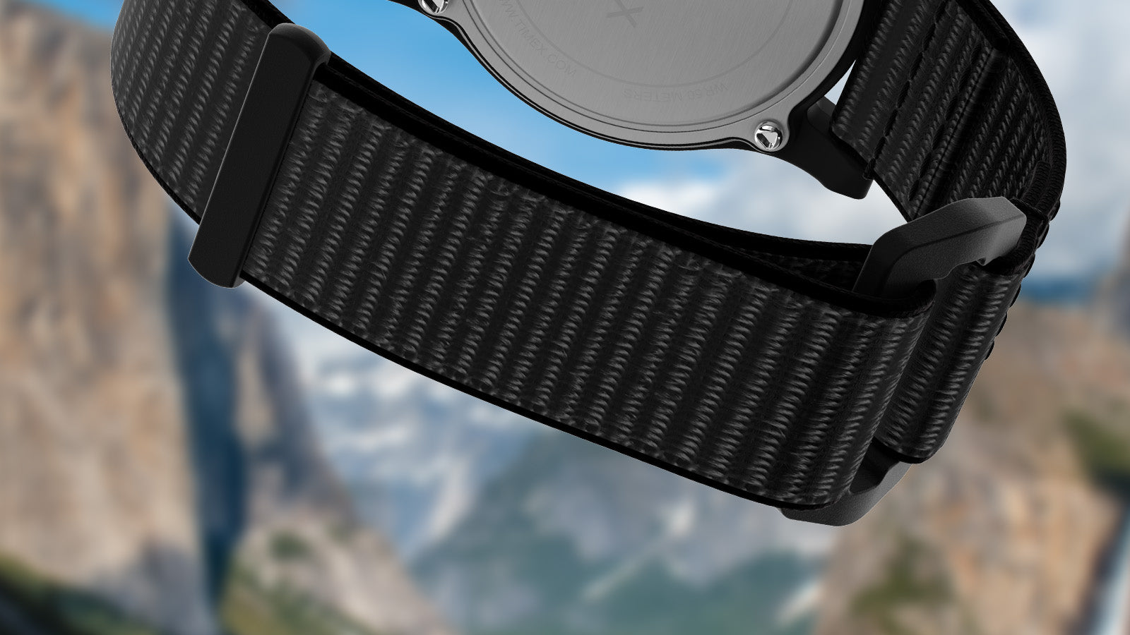 Detail of the black woven fabric strap and buckle of the Timex Yellowstone National Park watch, shown against a softly blurred mountain backdrop.