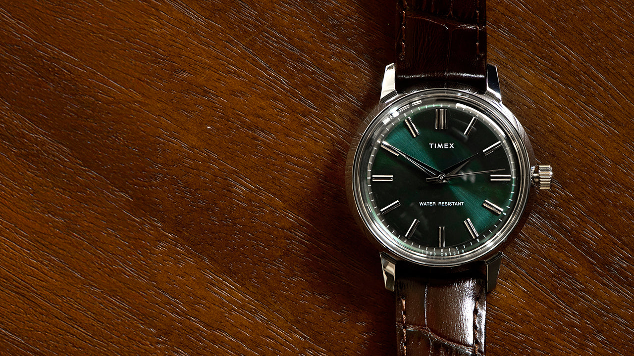 Todd Snyder x Timex Olive Marlin Automatic watch with a green sunray dial and brown leather strap, displayed flat on a textured wooden surface.