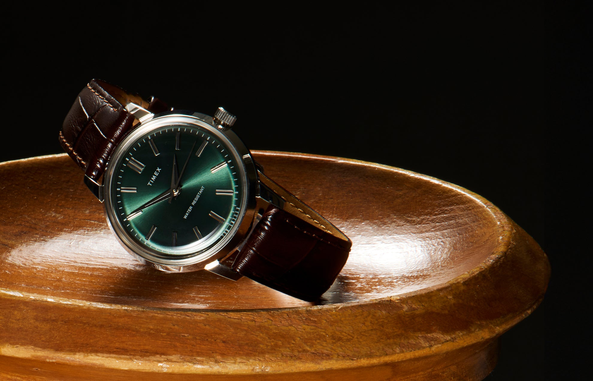 Todd Snyder x Timex Olive Marlin Automatic watch with a green dial resting at an angle on a polished wooden pedestal against a dark background.