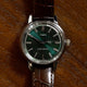 Front-facing view of the Todd Snyder x Timex Olive Marlin Automatic, showing its green dial, silver markers, and brown leather strap on a wood background.