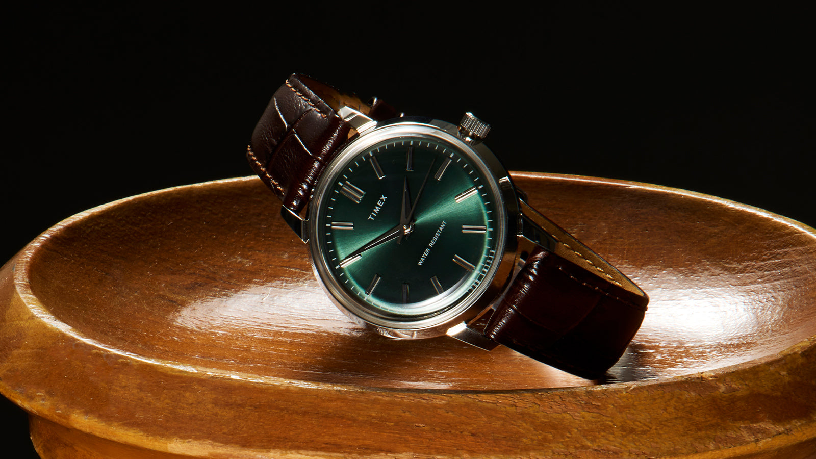 Todd Snyder x Timex Olive Marlin Automatic watch with a green dial resting at an angle on a polished wooden pedestal against a dark background.