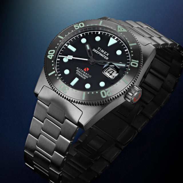 Timex Deepwater Reef 200 Titanium watch displayed at an angle, highlighting the black dial, aqua luminescent markers, titanium bezel, and matching titanium bracelet.