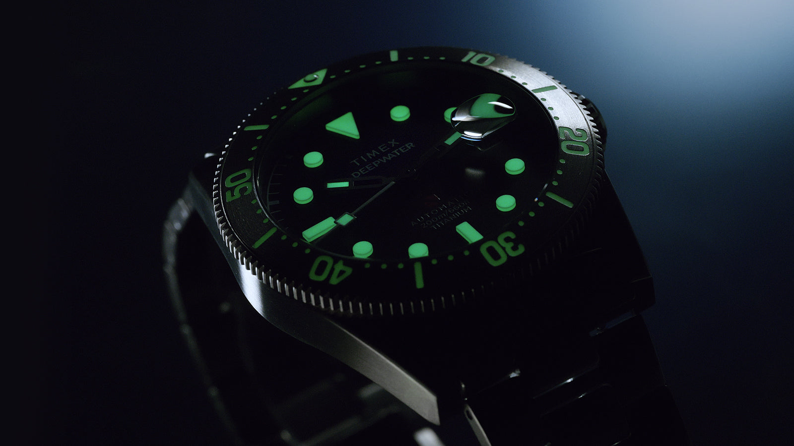 Timex Deepwater Reef 200 Titanium watch in low light with green luminescent hour markers, hands, and bezel indices glowing brightly.