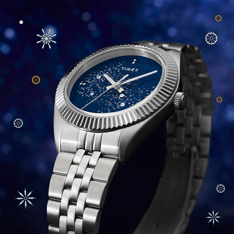 Timex Legacy Celestial watch with a stainless-steel case and bracelet, featuring a deep blue dial decorated with sparkling star-like details. The watch is shown against a cosmic-themed background with soft bokeh lights and small celestial graphic elements.