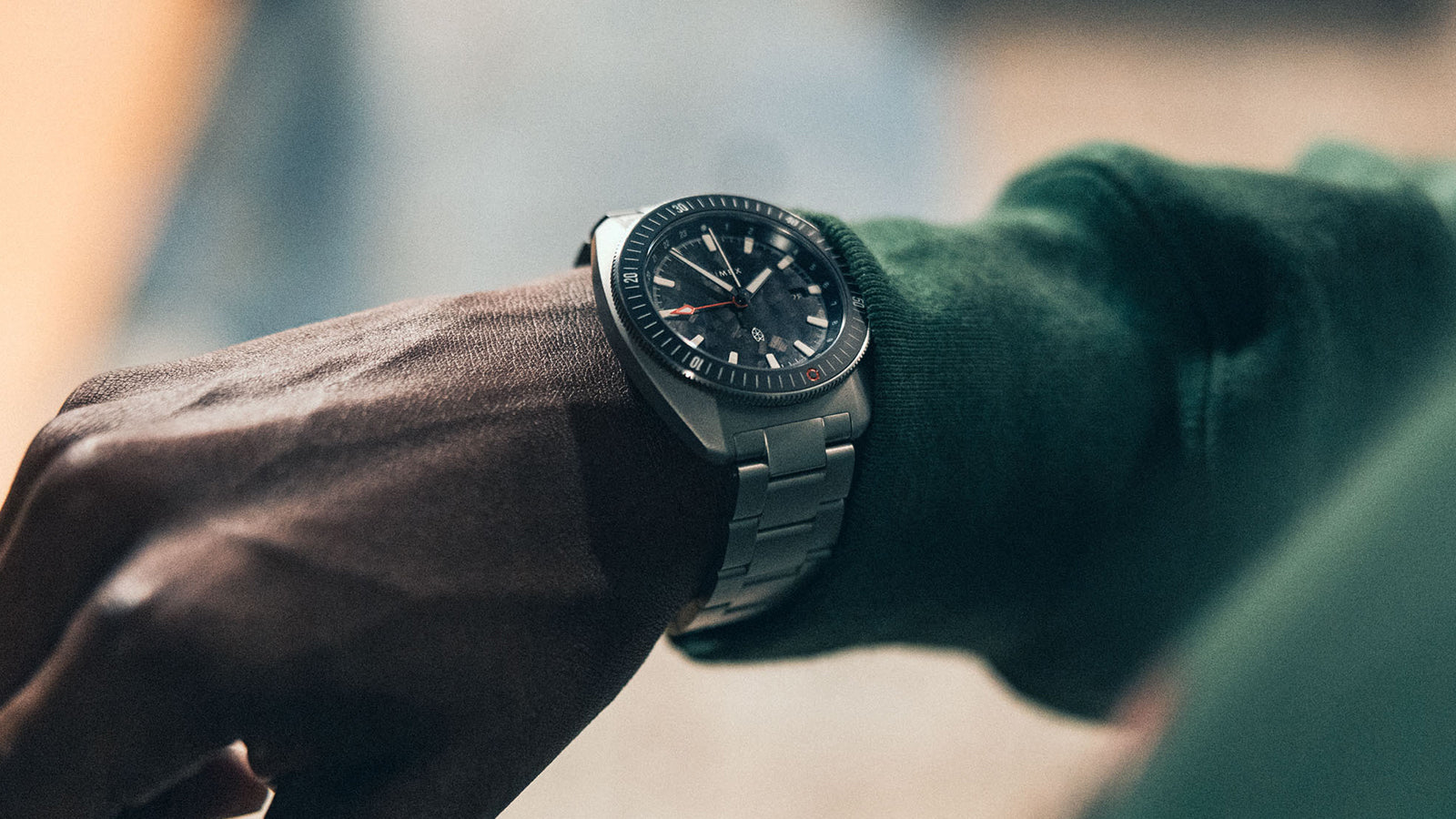 A close-up of a person wearing a Timex x James Brand wristwatch with a stainless steel bracelet and a black dial, shown on their left wrist against a blurred background. The person is dressed in a green long-sleeve shirt.